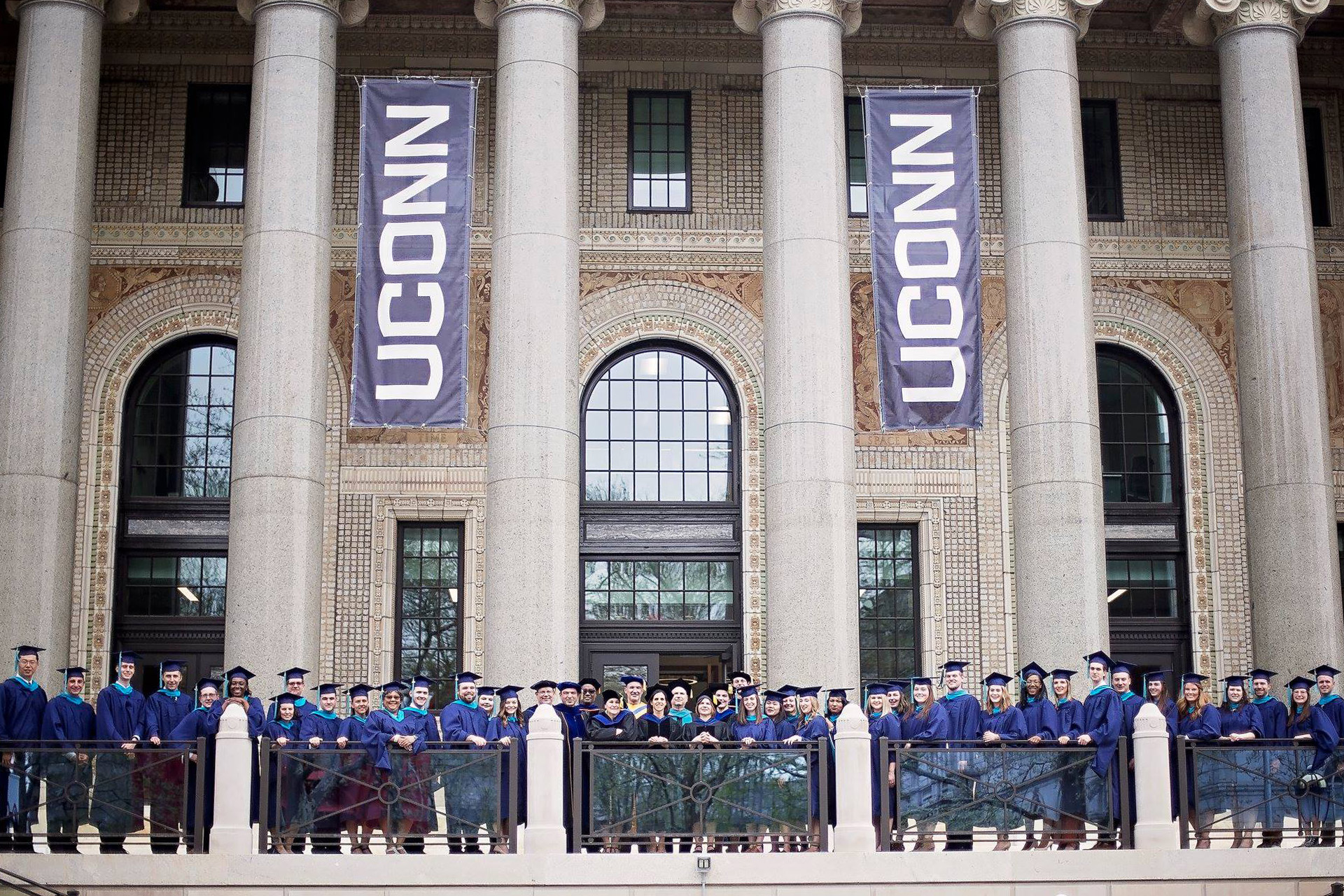 A large group of alumni and faculty in academic regalia lined up in front of the Hartford Times Building beneath two large UConn banners.